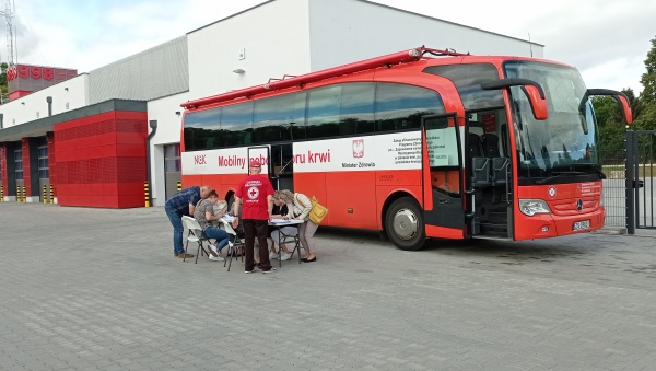 Na zdjęciu mobilny punkt poboru krwi (krwiobus), przed krwiobusem uczestnicy akcji wypełniają ankietę przy pomocy wolontariusza