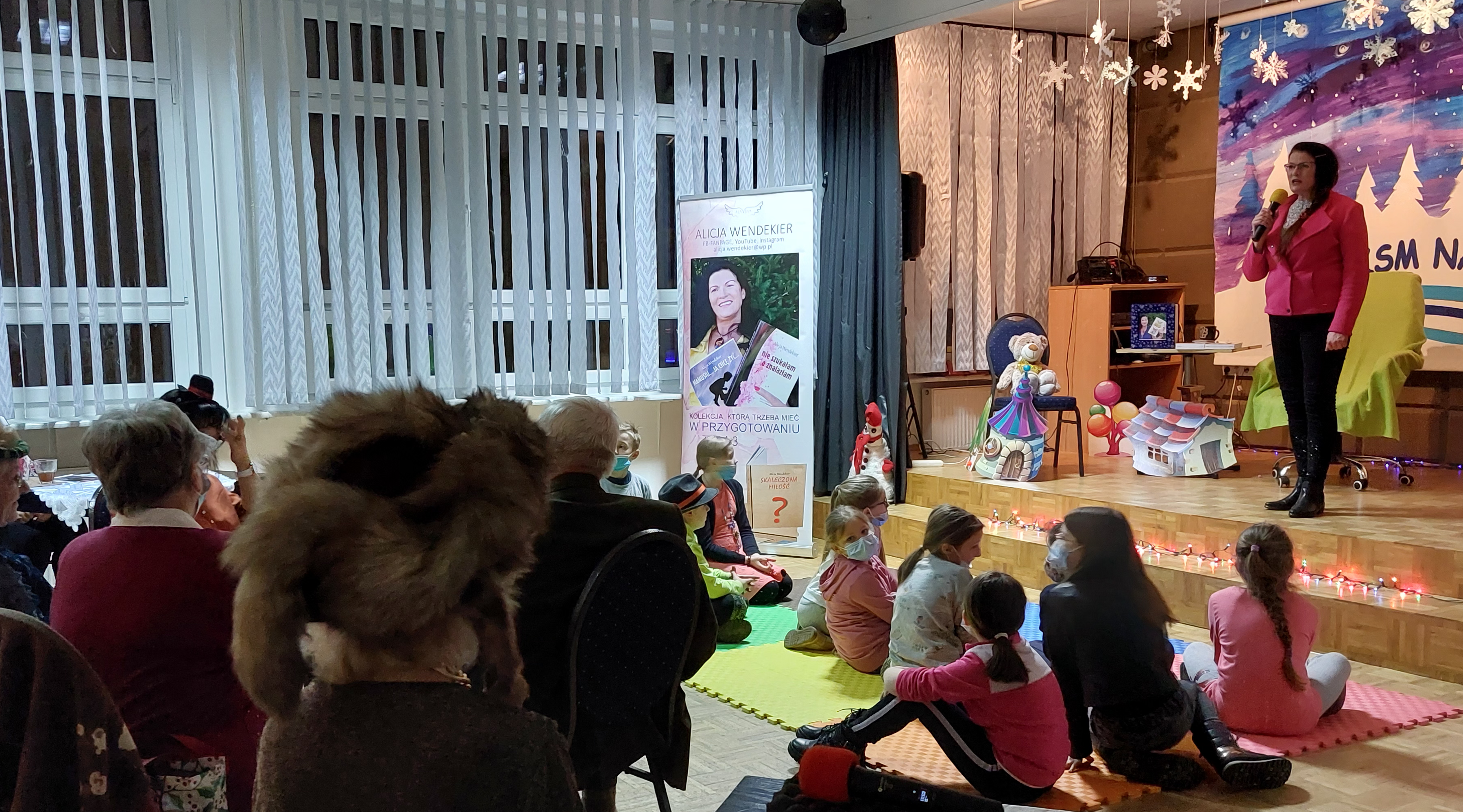 Pisarka stoi na scenie, seniorzy oraz dzieci wysłuchują jej przekazu motywacyjnego